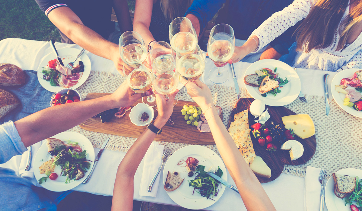 A group of people cheers with glasses of white wine over a table with food on plates, aerial view