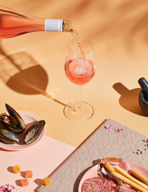 Product photography by the Brand Good Pair Days. A glass of rose wine is being poured. There's a pale orange background and plates of oysters and antipasto. There are deep shadows from the products.