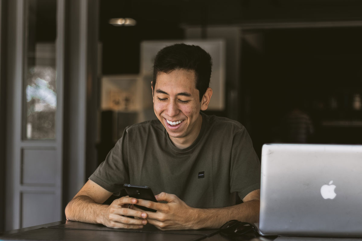 A man is smiling, looking at his mobile phone. He has a computer next to him.