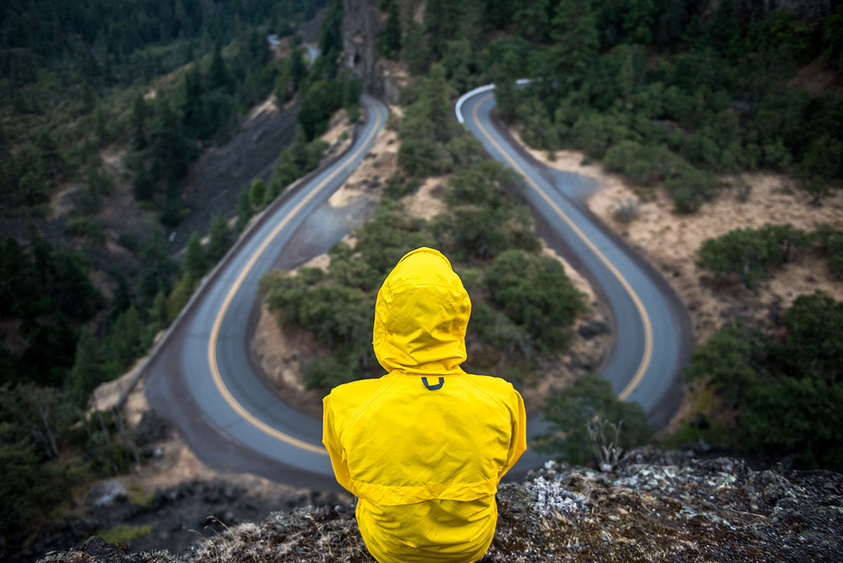 A person wearing a yellow raincoat sits with their back to the camera. They are looking at a winding road.