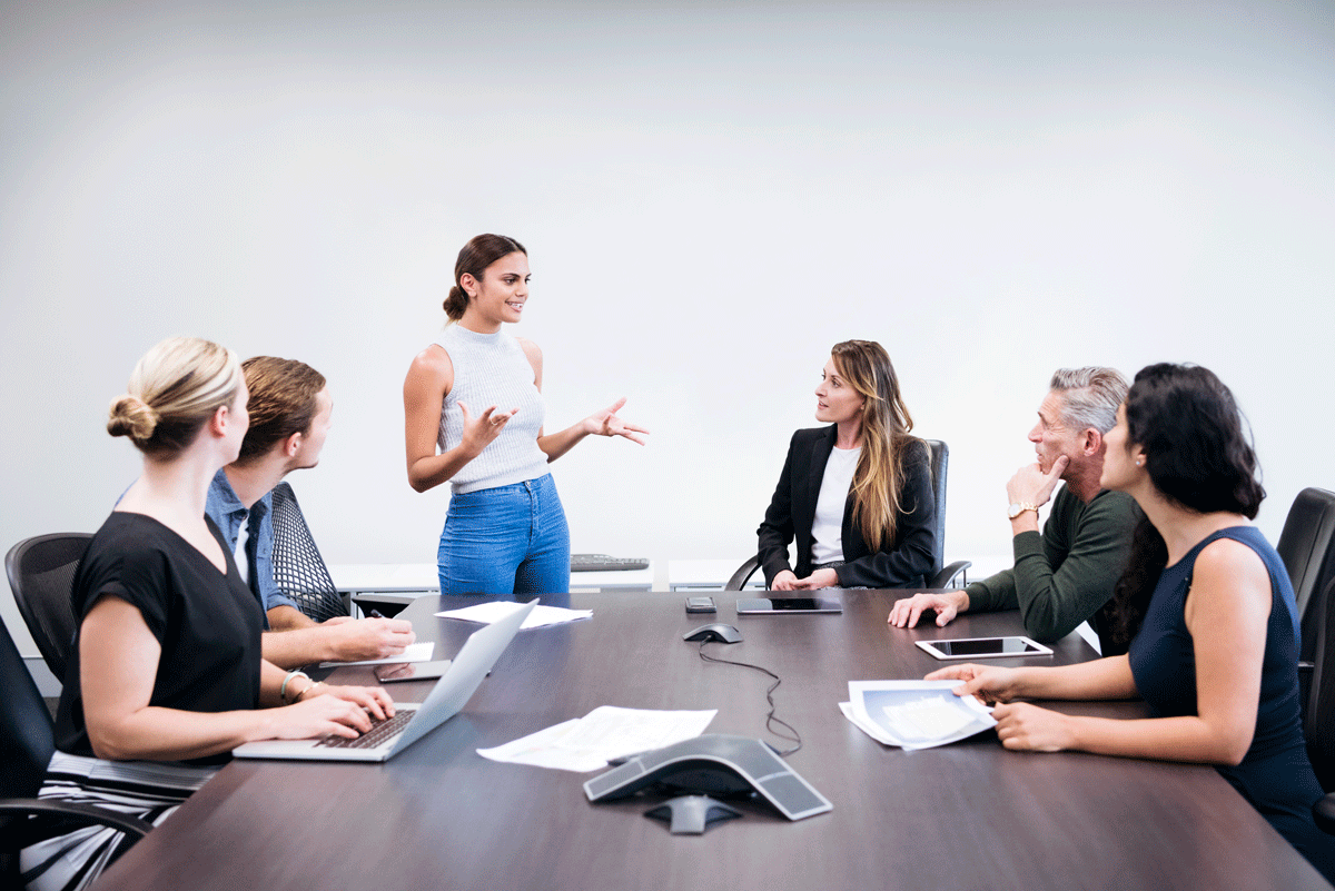A young woman stands in front of a conference room table of people, confidently speaking.