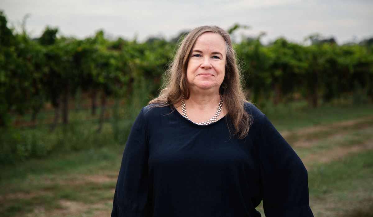A woman stands in front of a vineyard