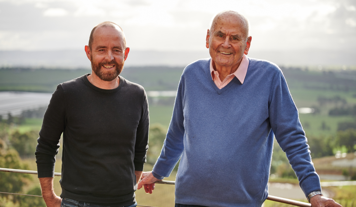 Two men smile for the camera; vineyards in the background