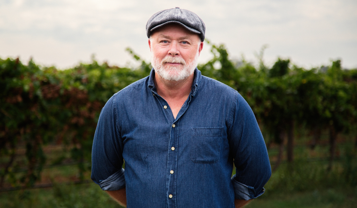 A man with a silver beard wearing a denim shirt and flat cap stands in front of a vineyard