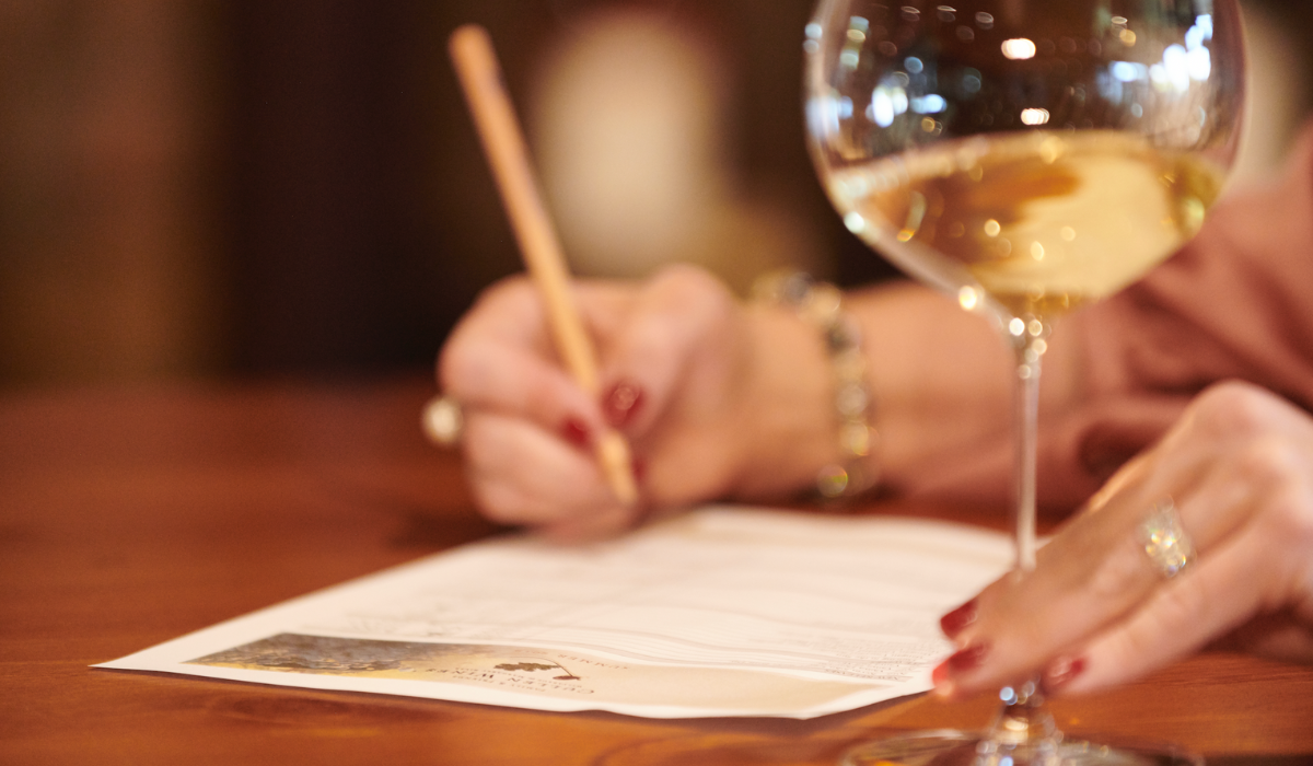 Close up of a glass of white wine and someone's hand taking notes