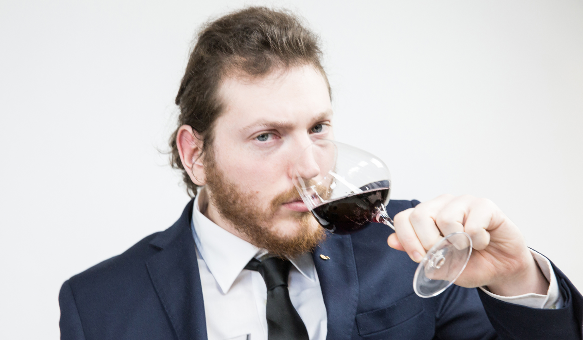 A man with a beard and a ponytail smells a glass of red wine and stares into the camera