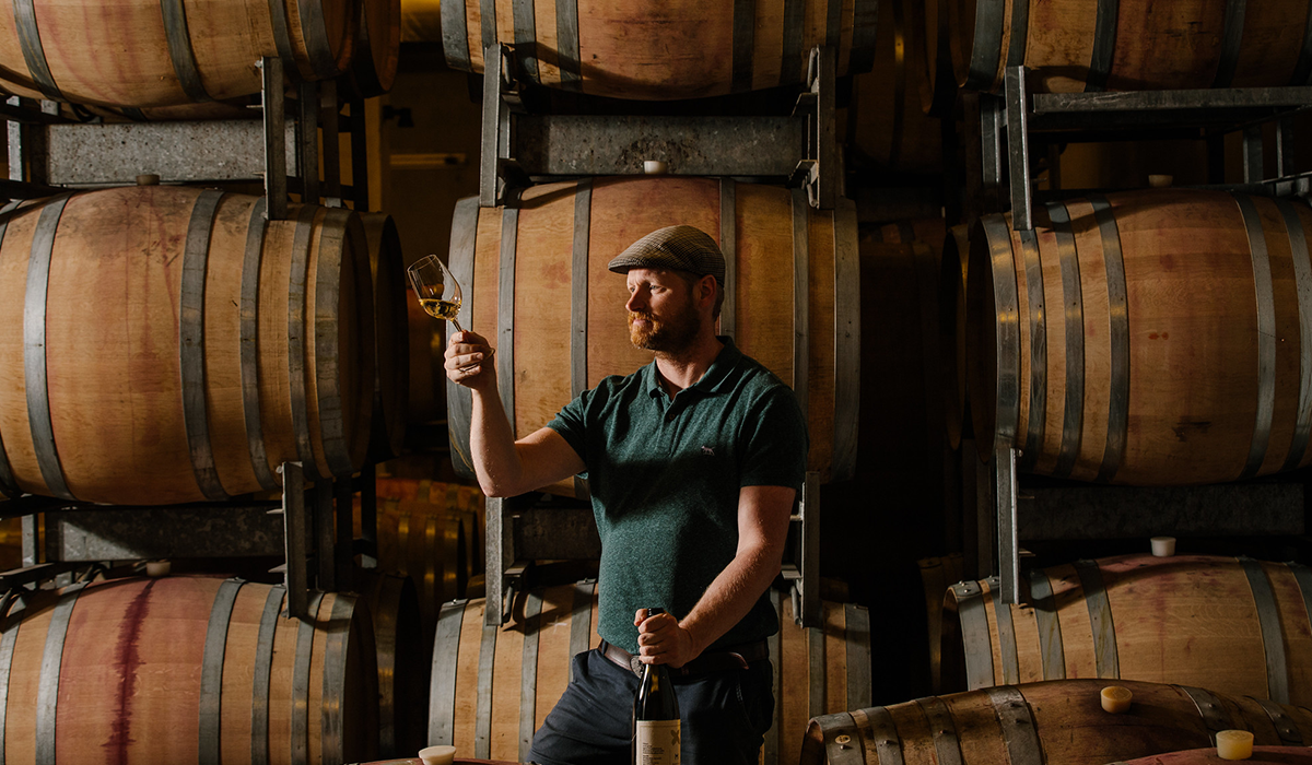  Conor van der Reest holding up a glass of white wine with wine barrels behind him