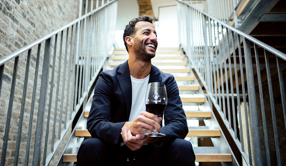 Daniel Ricciardo sitting on a set of stairs holding a glass of red wine with his head turned to the right, and smiling