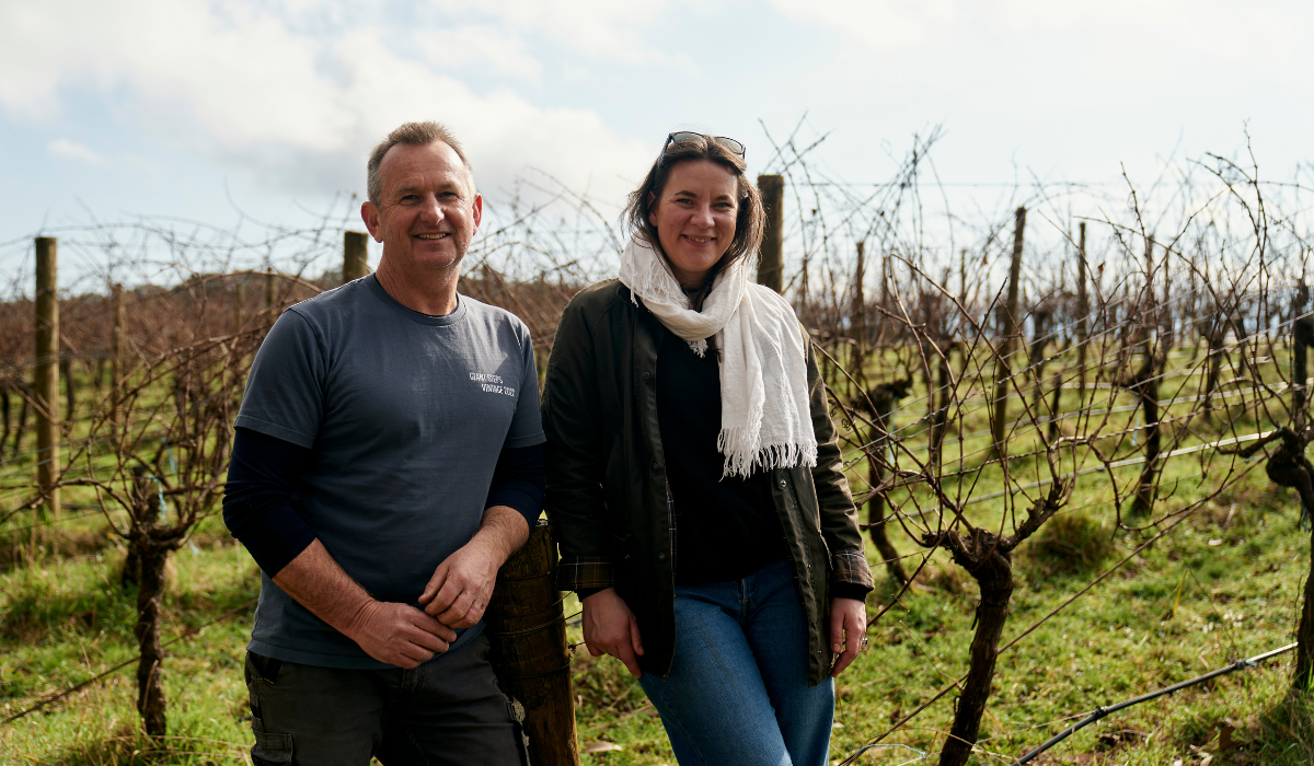 A man and woman pose in front of a vineyard