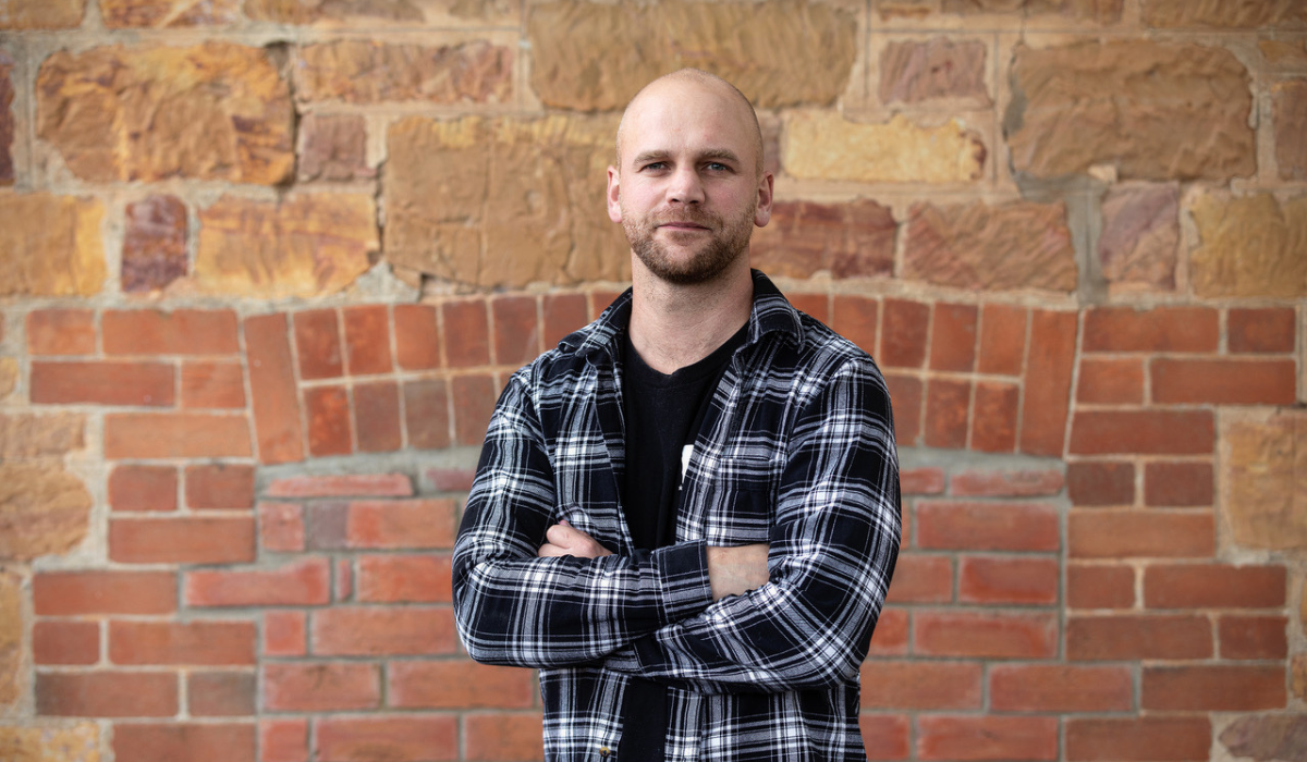 A man in a plaid shirt poses in front of a brick wall