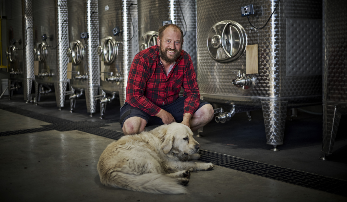 A man in a red shirt poses with his dog in front of fermentation tanks