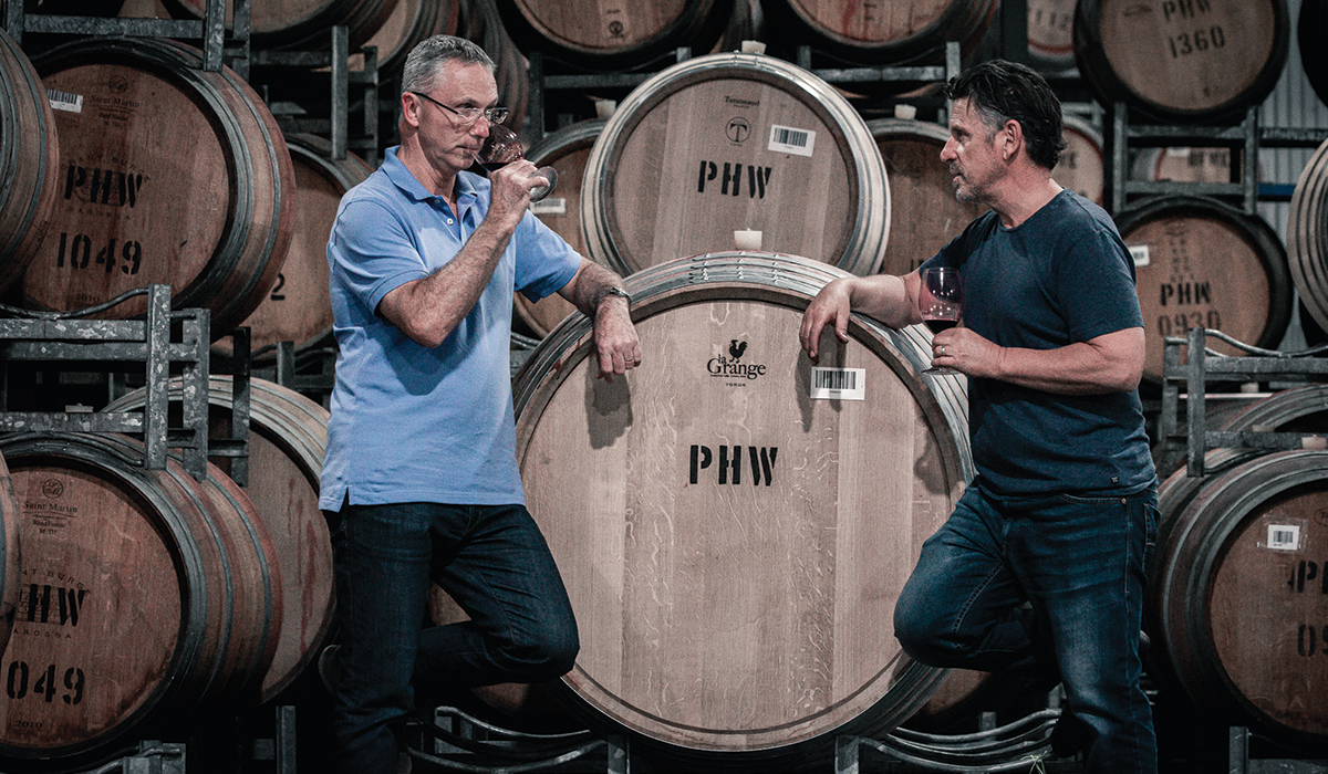 Two men drinking wine in front of barrels