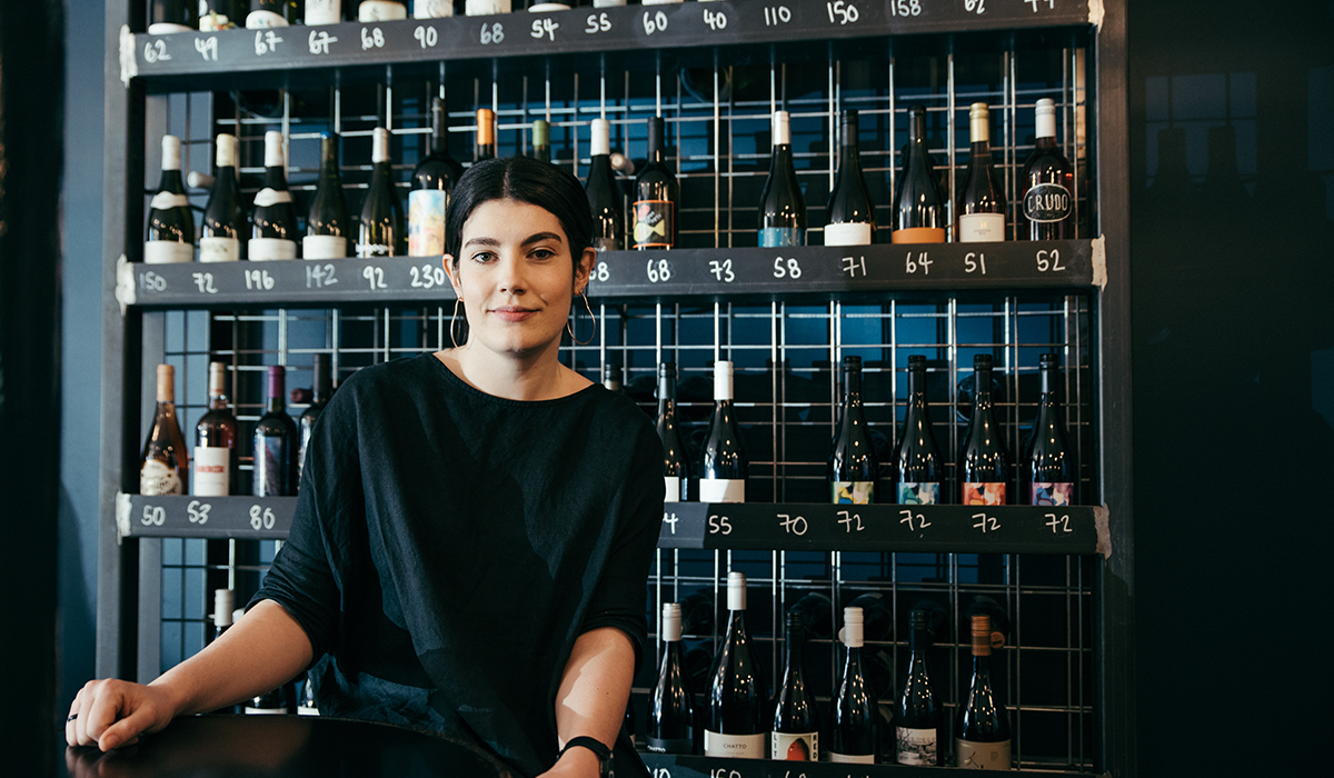 Alice sitting down, wearing a black top with a rack of wines behind her.