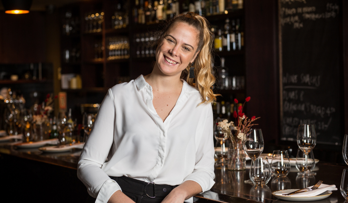 A woman wearing a white shirt stands in front of a bar