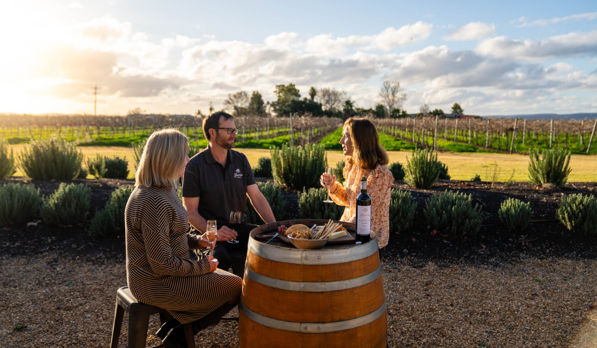 Three people sit around a wine barrel sipping wine and eating cheese